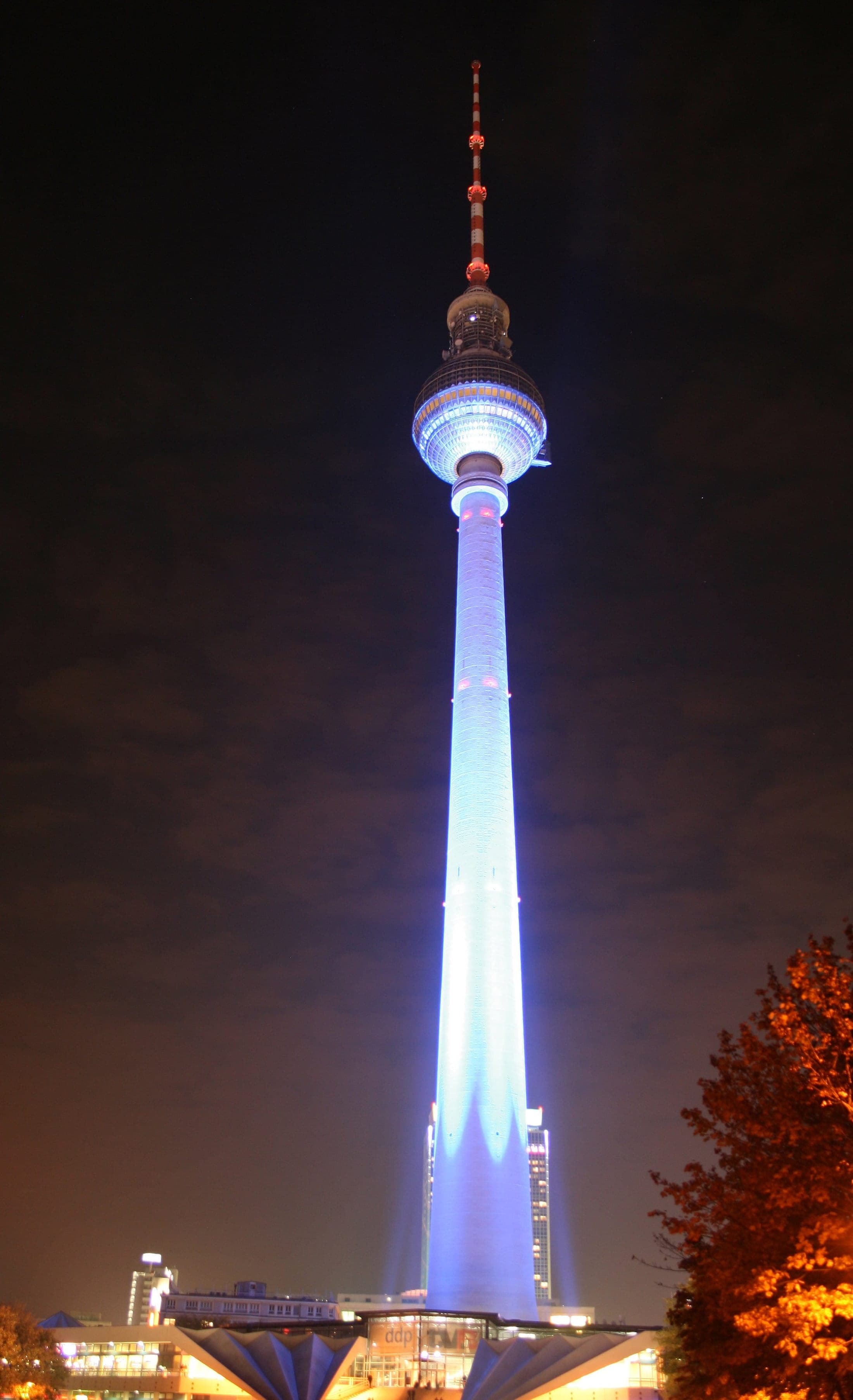 The Berlin television tower at night during the Festival of Lights, its sphere lit in bands of colour above the dark city.