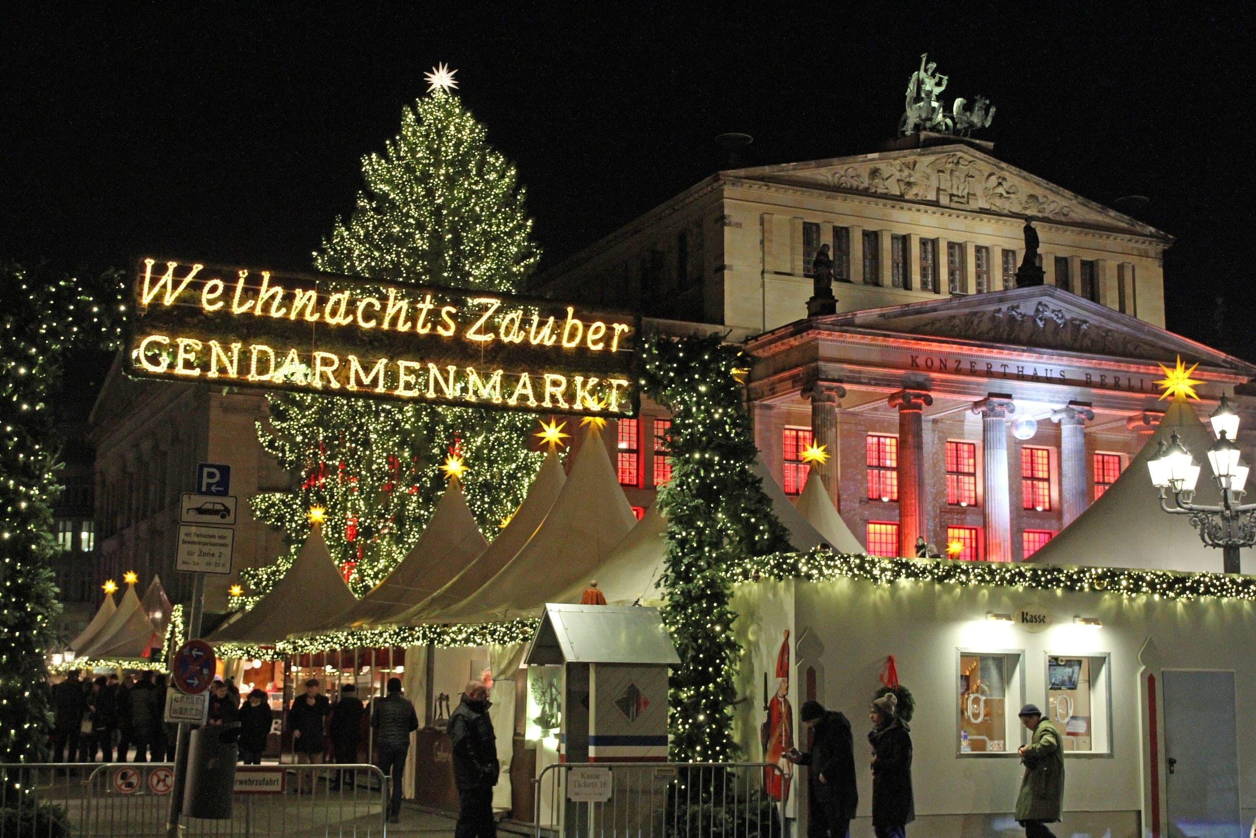 The Christmas market on Gendarmenmarkt after dark, rows of white peaked stall roofs strung with lights in front of the floodlit domed cathedral.