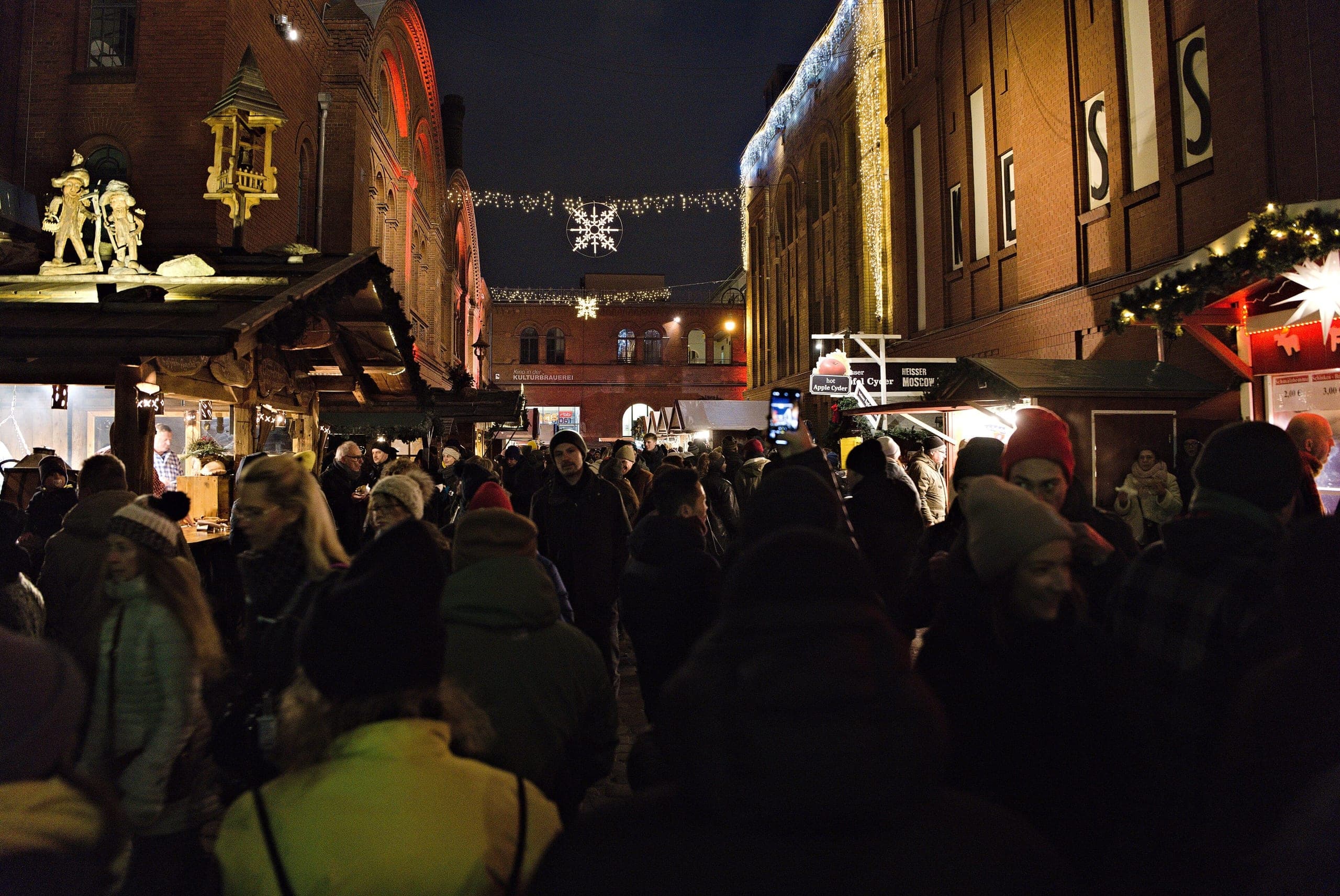 The Christmas market in the Kulturbrauerei courtyard at night, wooden stalls and strings of warm lights between tall red-brick industrial buildings.