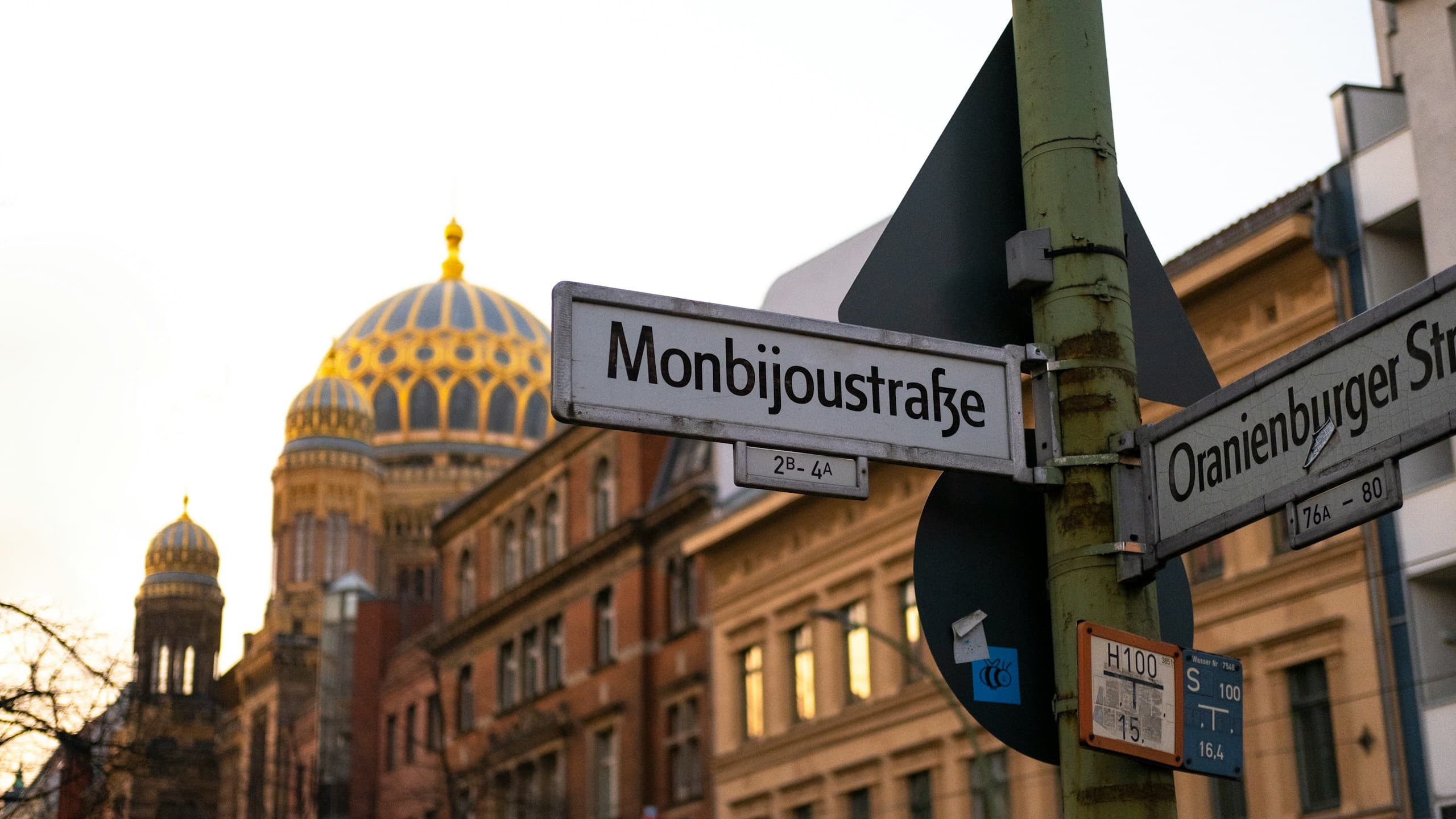 A street sign at the corner of Monbijoustraße and Oranienburger Straße in Mitte at dusk, with the gold dome of the New Synagogue behind it.