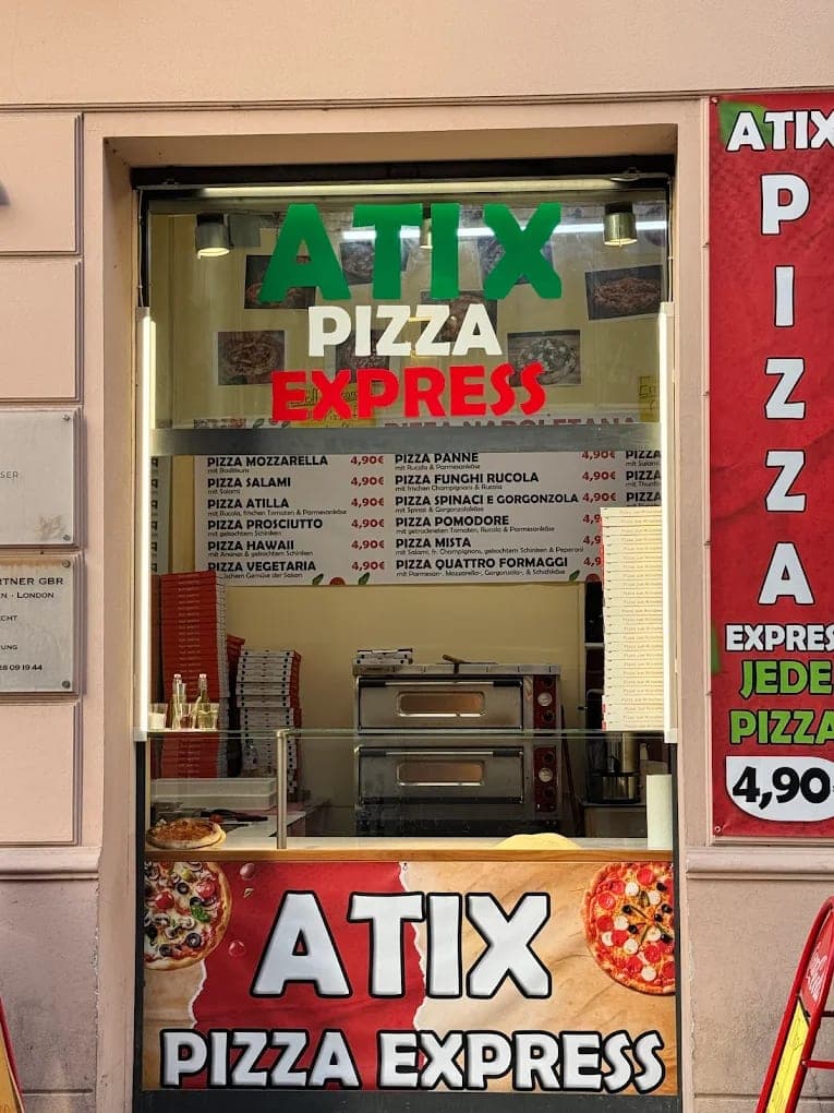 The shopfront serving window, with green and red “ATIX Pizza Express” signage above a printed menu board and a pizza oven visible inside.