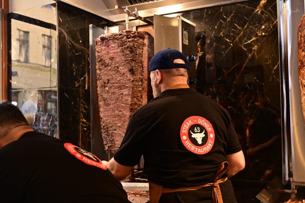 A cook in a black shirt, seen from behind, holding a red tray beside a tall vertical rotating spit against a dark marble wall.