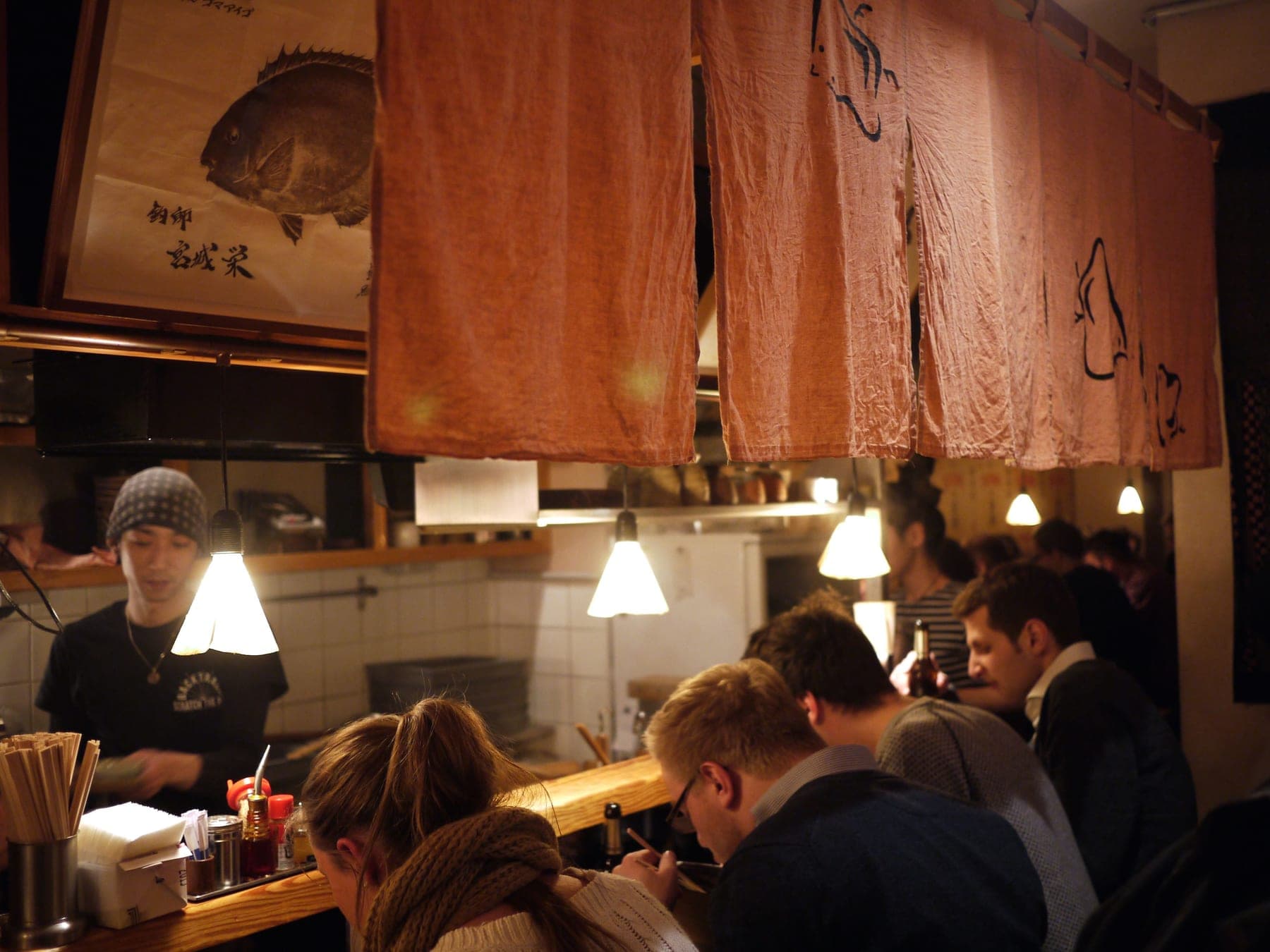 Guests seated along a wooden counter under low pendant lamps, with cooks working in the open kitchen behind and rust-coloured fabric curtains hanging above.