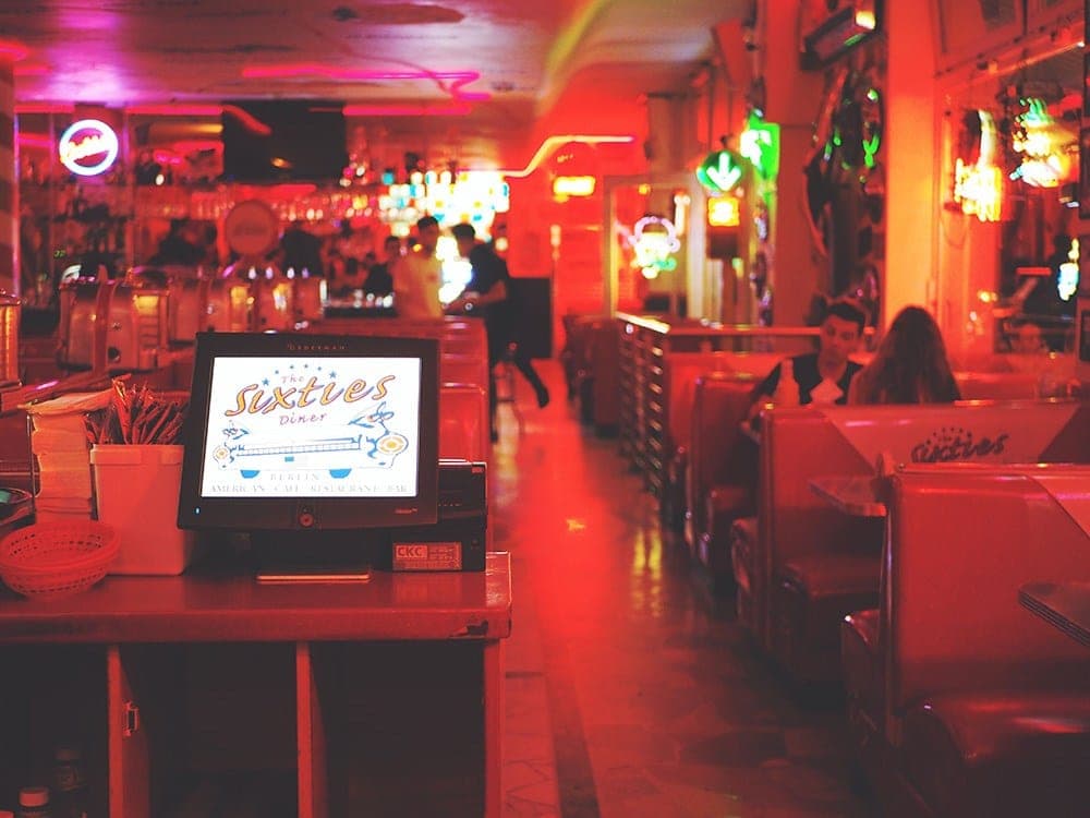 The diner’s interior under red neon light, with red booth seating along one wall and an illuminated menu screen reading “The Sixties Diner” on the counter.
