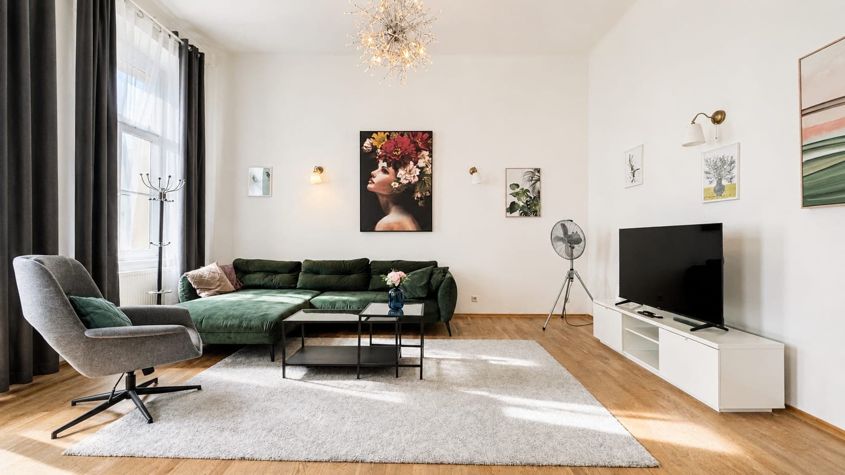 A tall Altbau living room with a green velvet corner sofa, a grey swivel armchair and a starburst chandelier, beside a full-height window in charcoal curtains.
