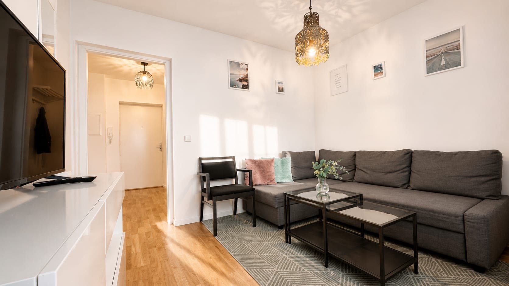 A pale, softly lit room with a grey corner sofa, pink and mint cushions, coastal photographs on white walls and a pierced brass pendant throwing patterns across the ceiling.