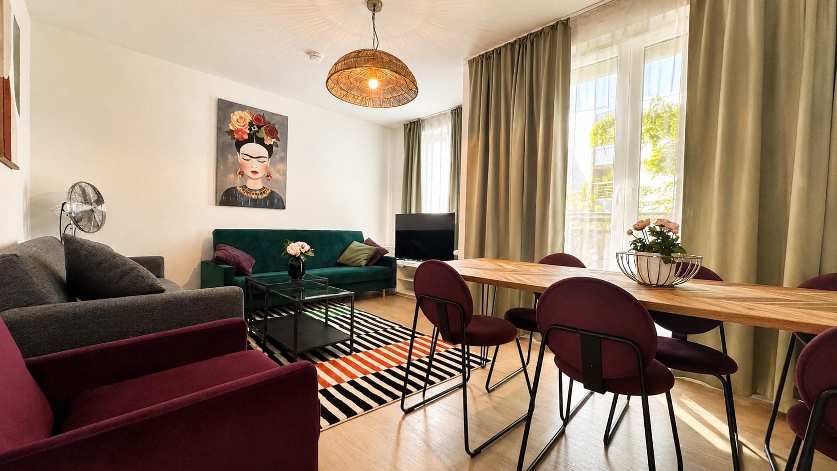 An open living and dining room with a dark green velvet sofa, a striped rug and a herringbone table under a woven pendant lamp.