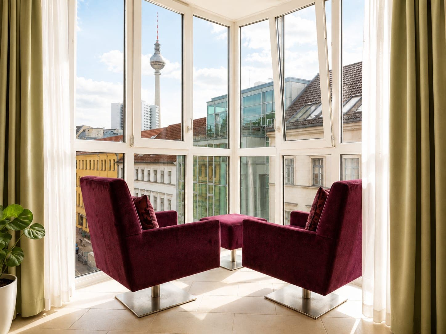 Two deep red velvet armchairs and a matching footstool in a corner window, with Berlin’s television tower rising over the rooftops beyond.