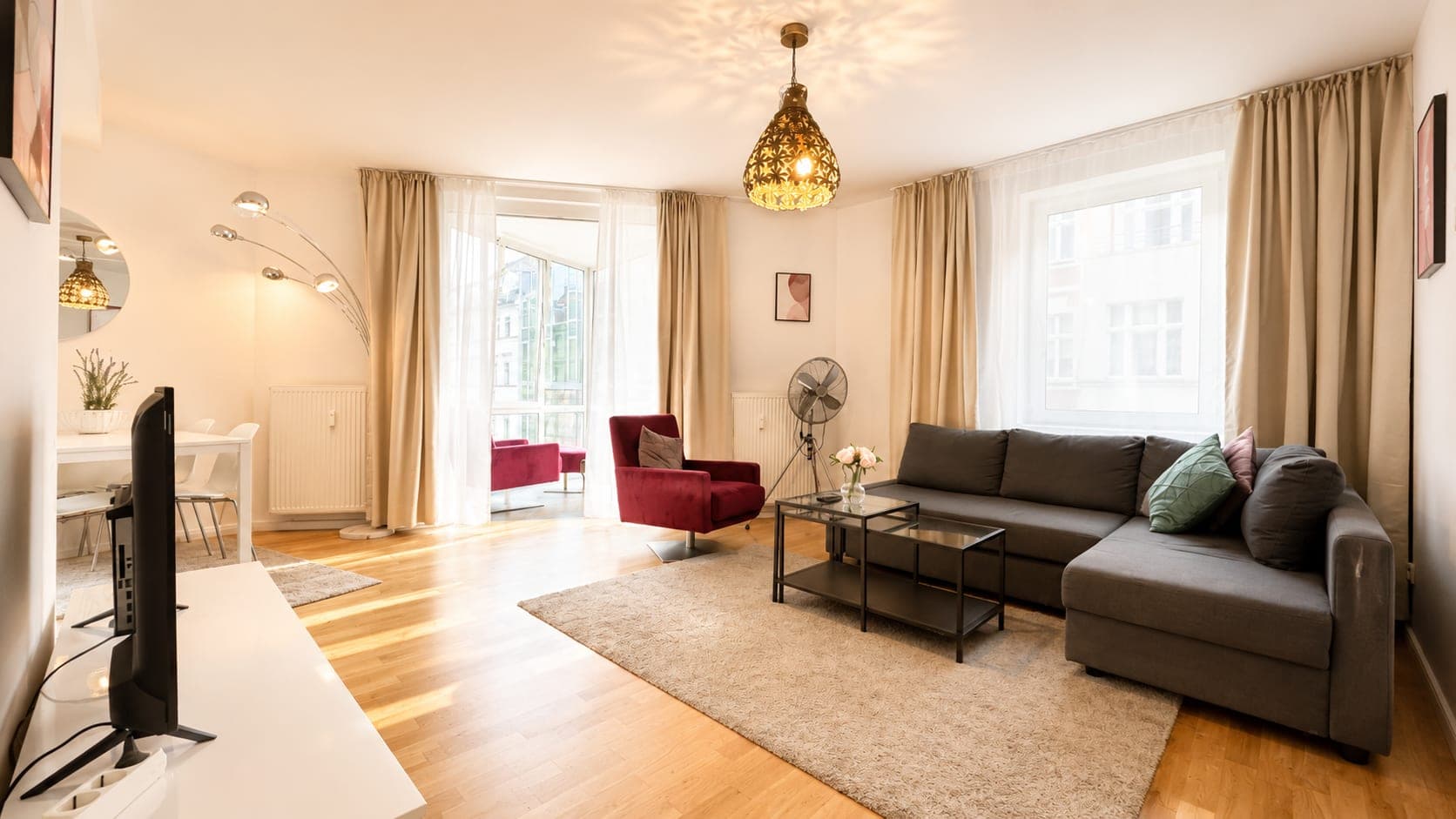 A living room in warm lamplight, with a grey corner sofa, a red velvet armchair, a patterned brass pendant lamp, and a balcony door beside a window looking onto the building opposite.